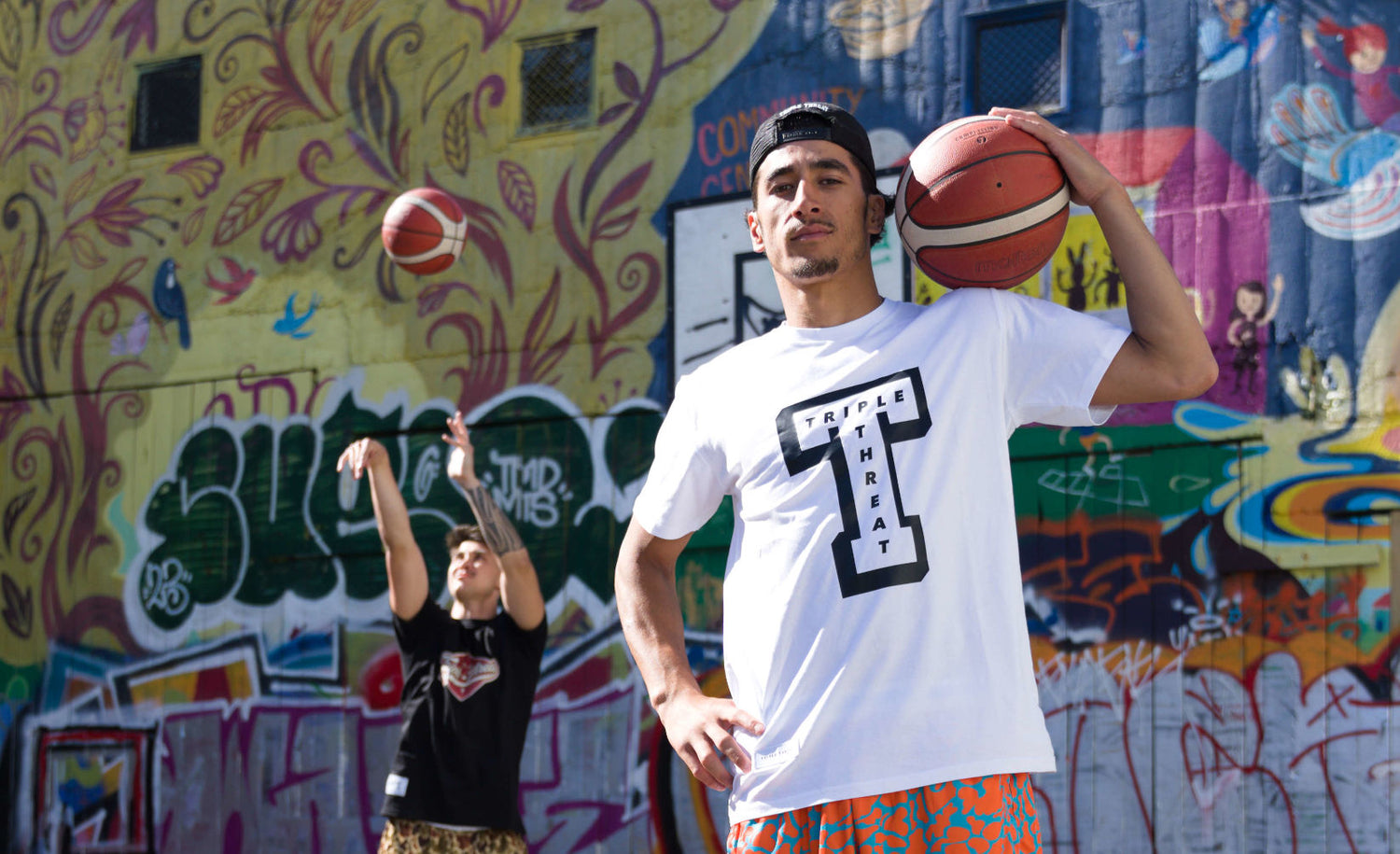 Two models wearing Triple Threat clothing with basketballs with a graffiti wall background