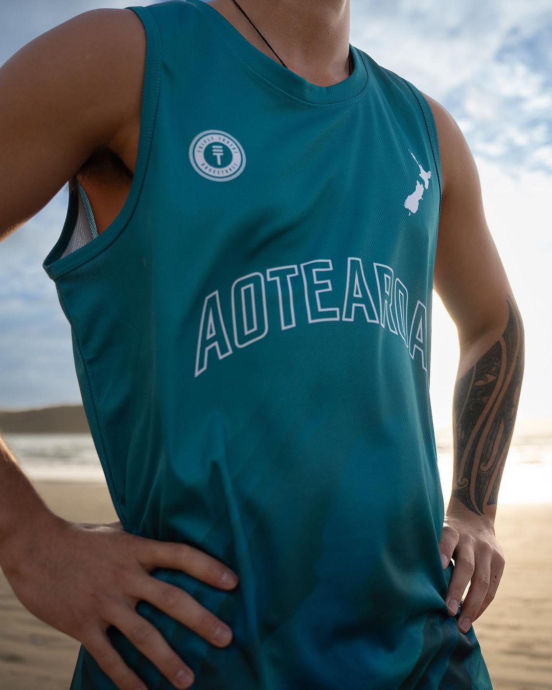 Man wearing teal paua basketball singlet with Paua design, New Zealand map, and Triple Threat logo, standing on the beach and the text 'Aotearoa' in white in the center