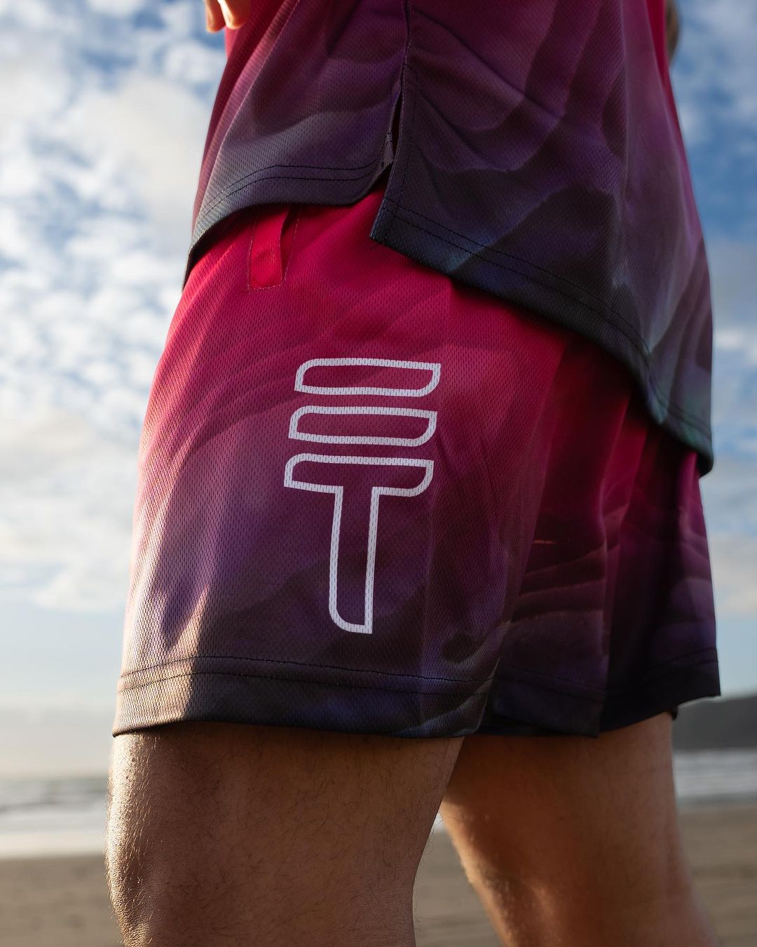 Pink basketball shorts with a Paua design at the bottom and a large white Triple Threat logo on the side being worn by man on the beach in NZ
