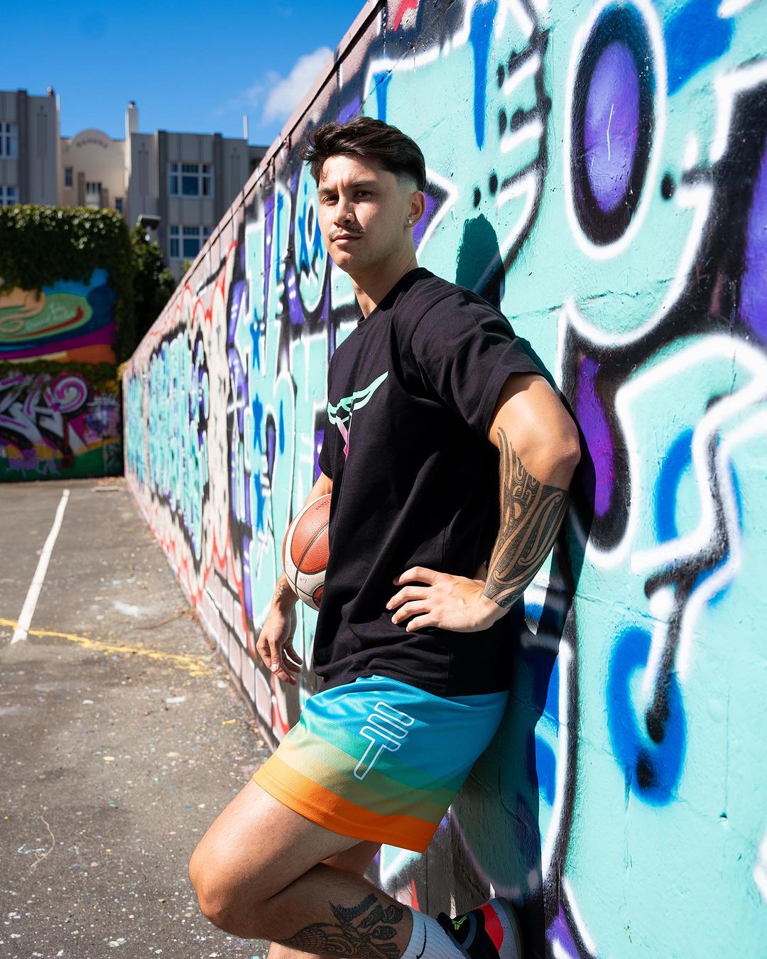 model wearing Triple Threat T-shirt and buffalo basketball shirts, leaning against a graffiti wall holding a basketball