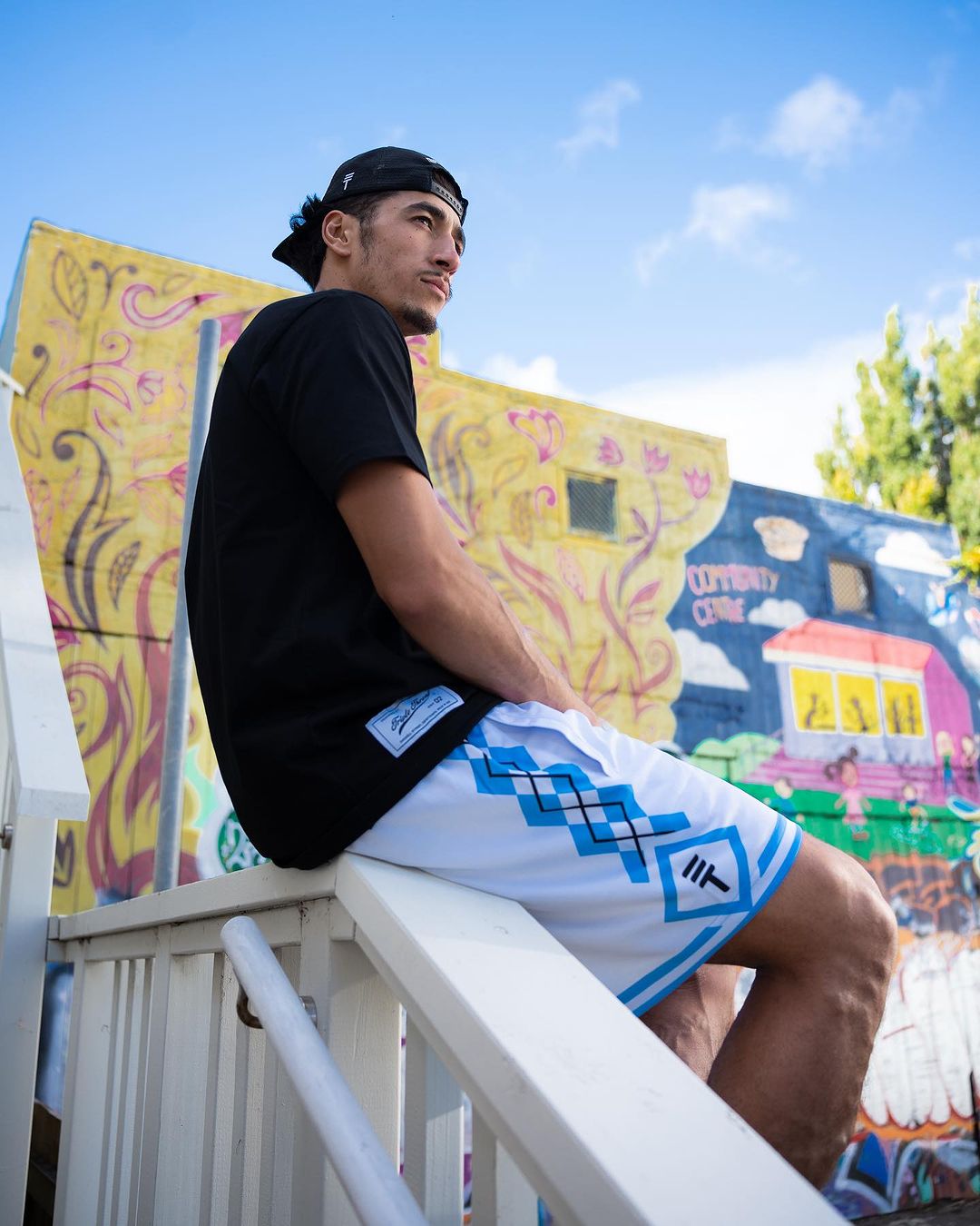 Man wearing white basketball shorts with light blue diamond pattern, elastic waistband, and the Triple Threat logo, sitting on a railing in front of a colorful mural.