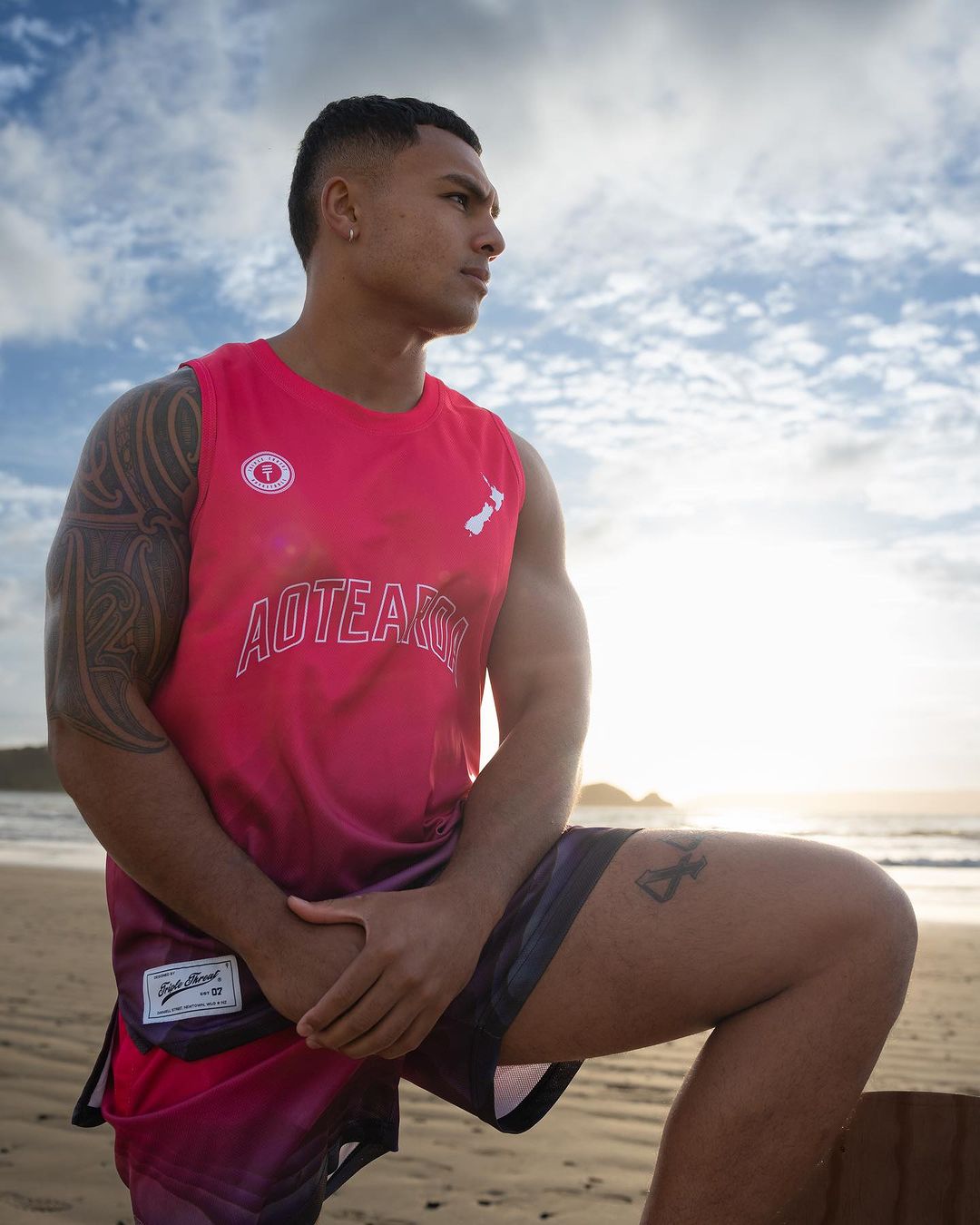 Man wearing pink Aotearoa basketball singlet with paua design and New Zealand map, and Triple Threat logo kneeling on the beach