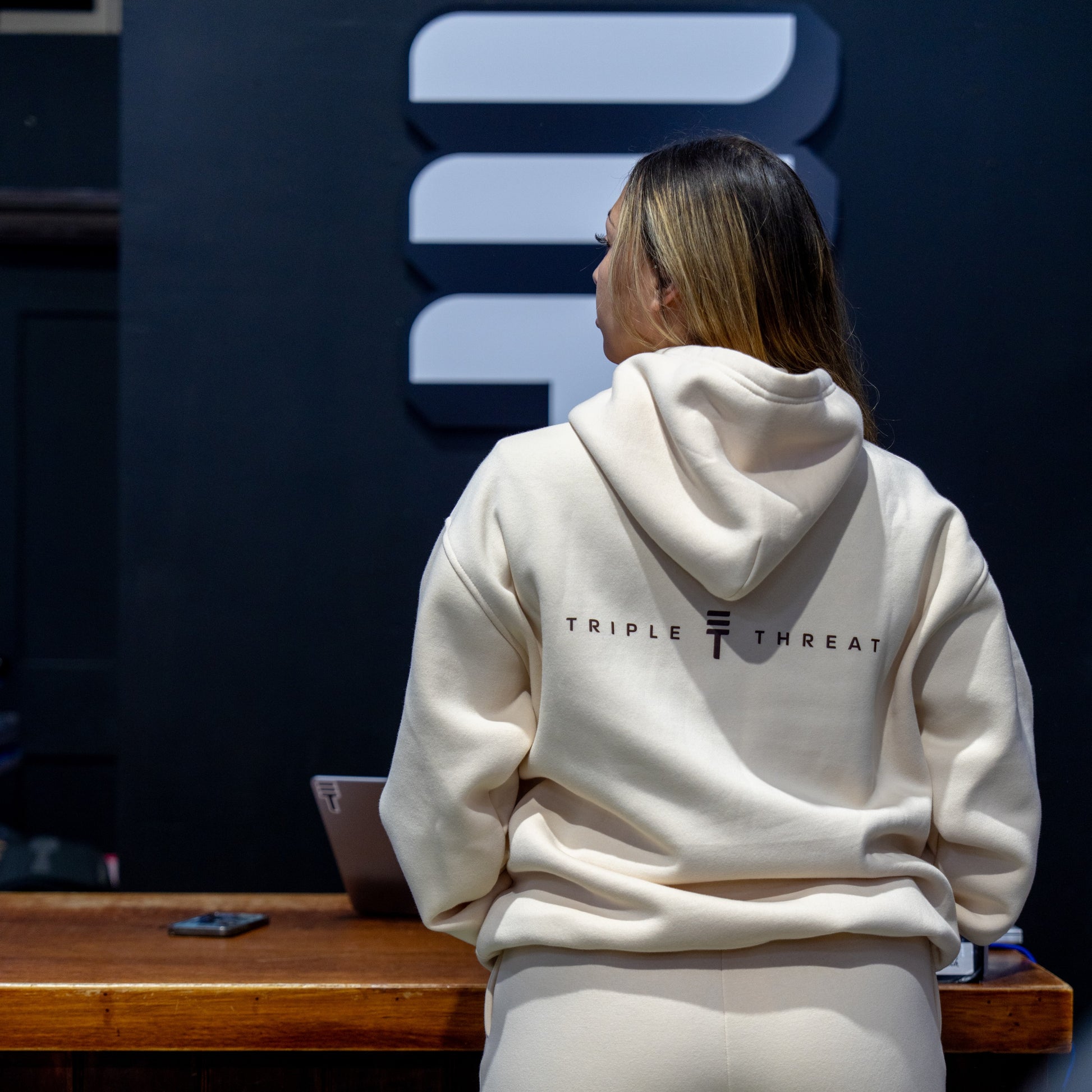 Model wearing a cream hoodie with 'Triple Threat' branding, standing in front of a dark background.