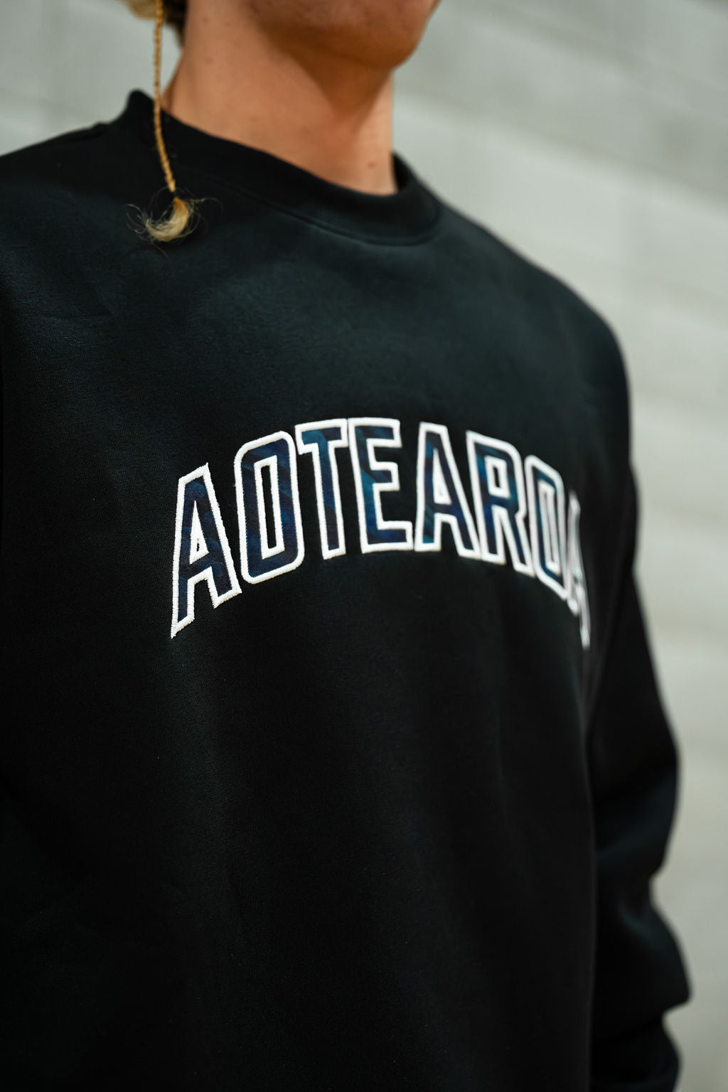 Black crewneck sweatshirt with 'Aotearoa' text worn by a person, blurred background