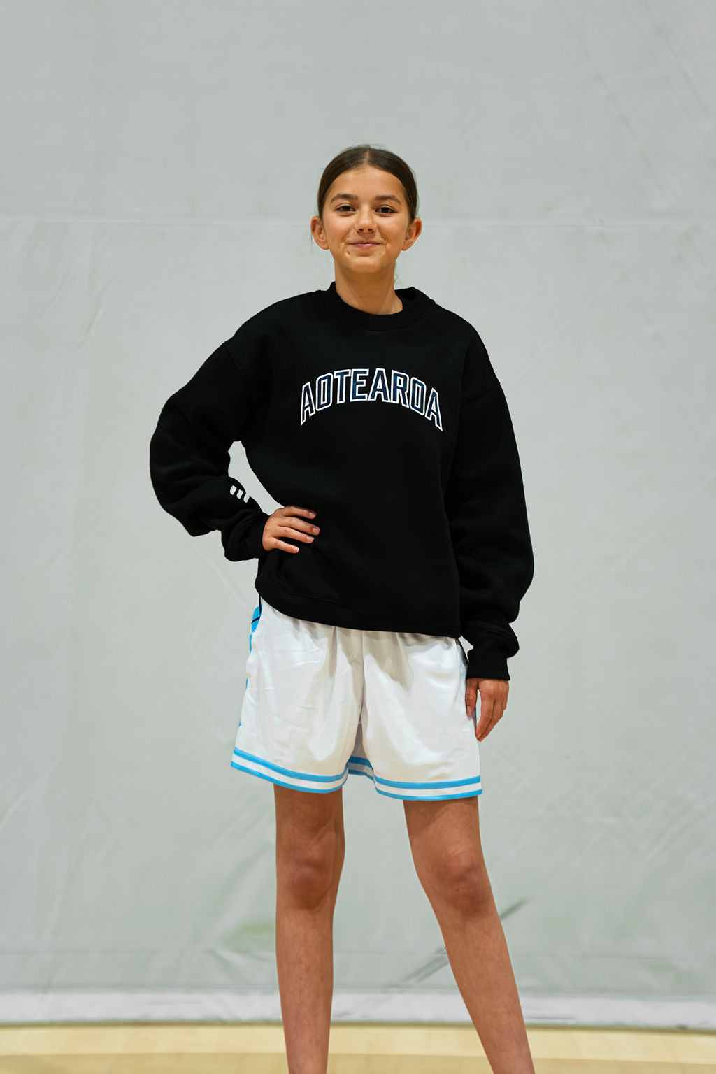 Person wearing a black kids sweatshirt with 'Aotearoa' text and white shorts with blue stripes on a plain background
