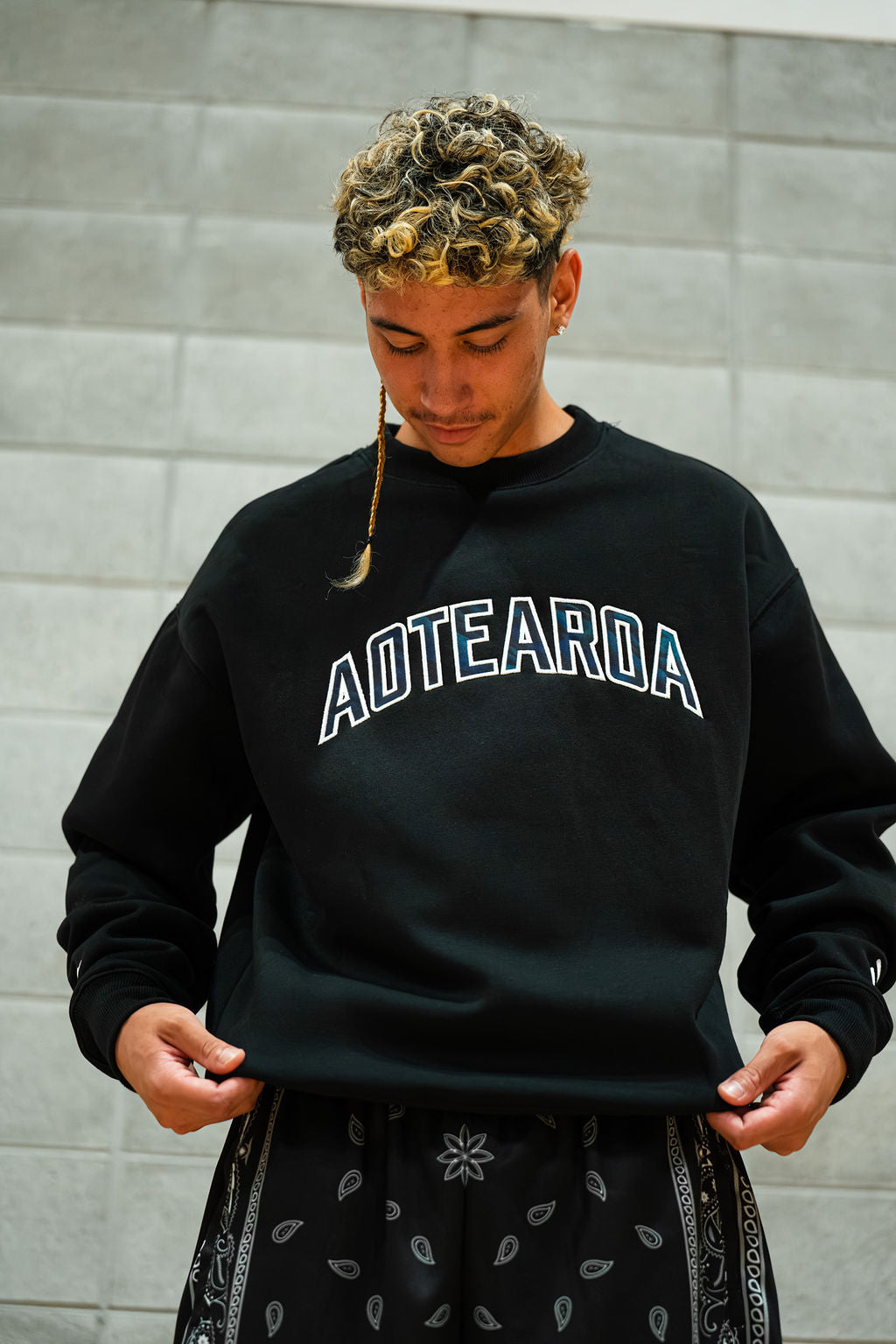 Person wearing a black sweatshirt with 'AOTEAROA' text against a grey brick wall.