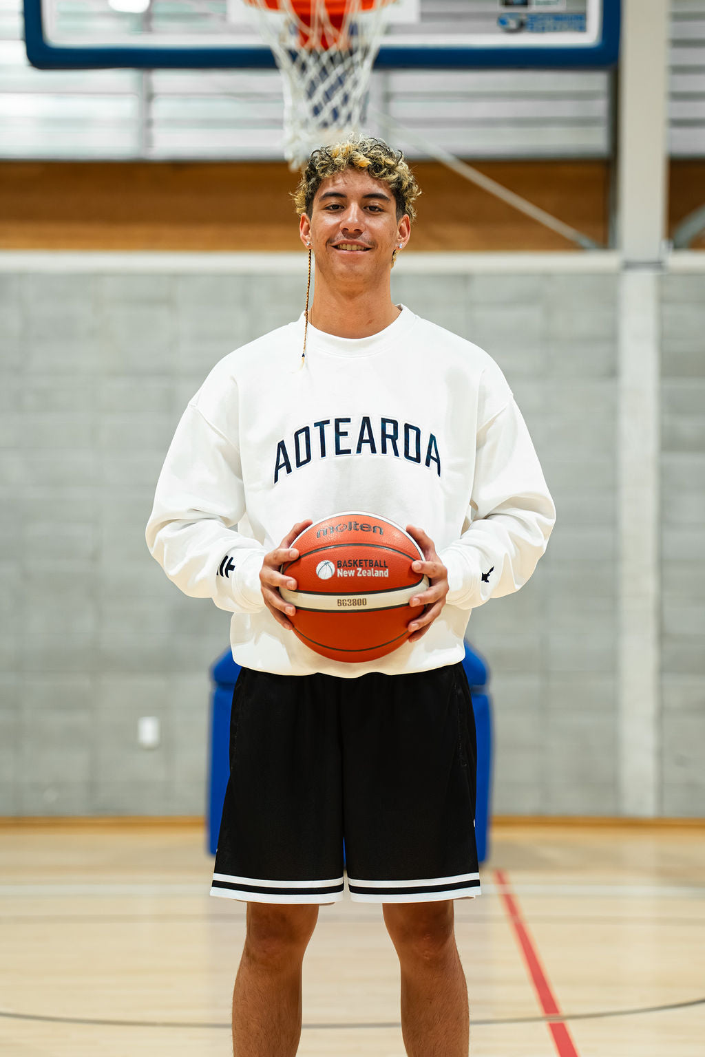 Person holding a basketball in a gymnasium wearing a white 'Aotearoa' sweatshirt.