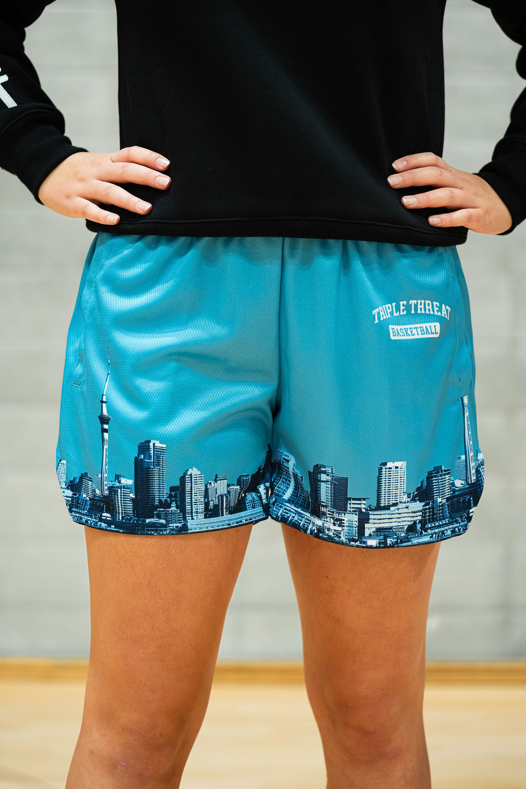 Person wearing teal blue basketball shorts with Auckland cityscape design and black top on a neutral background