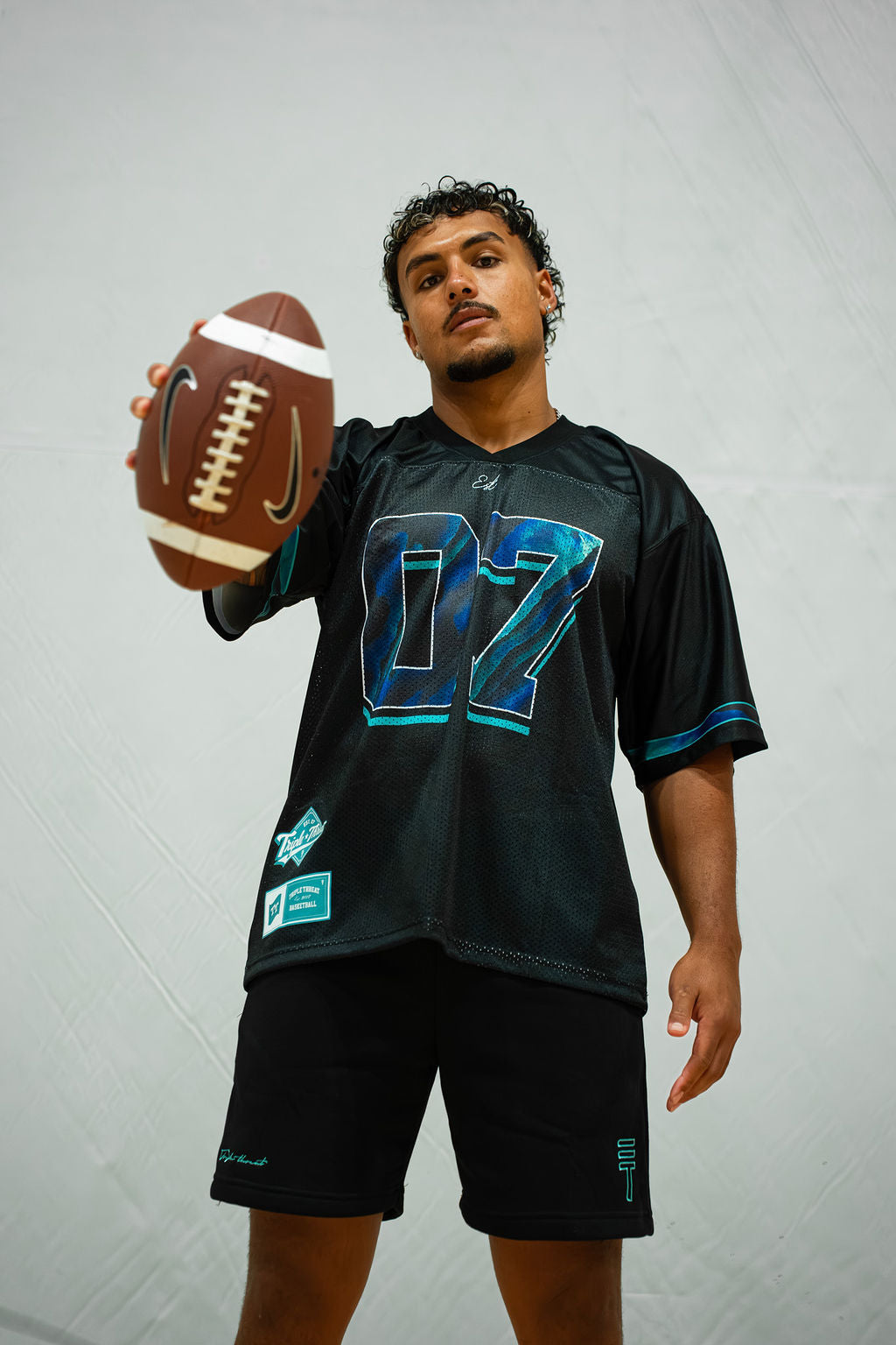 Person wearing black shorts and a black sports jersey with number 07 holding a football against a grey background