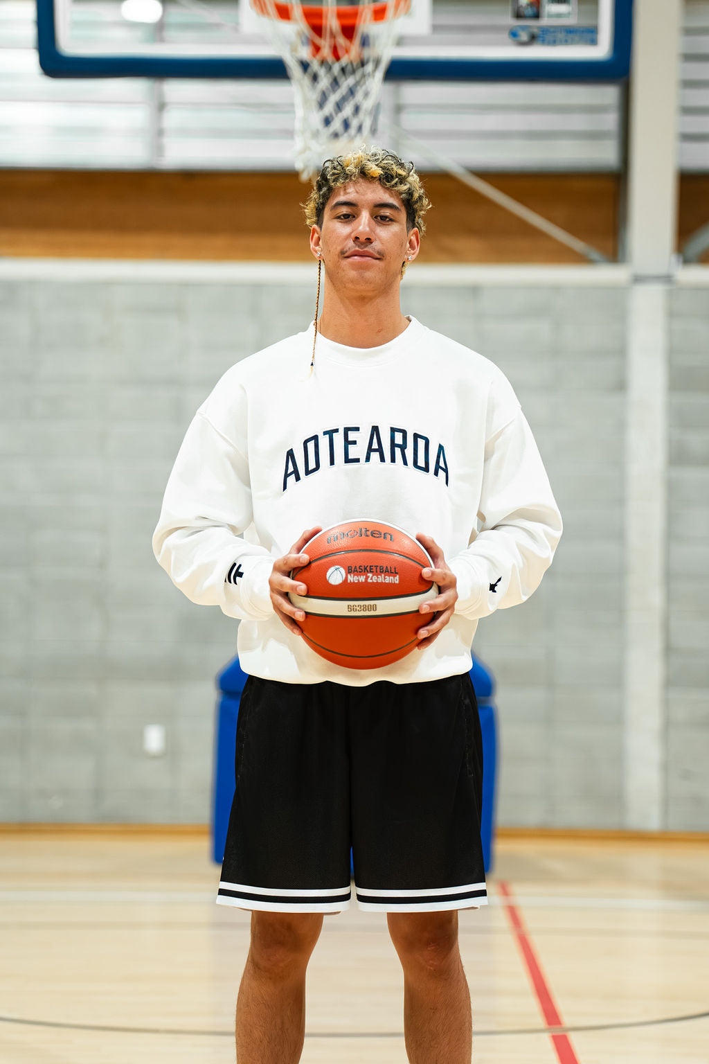 Person holding a basketball in black basketball shorts and white sweatshirt on an indoor basketball court