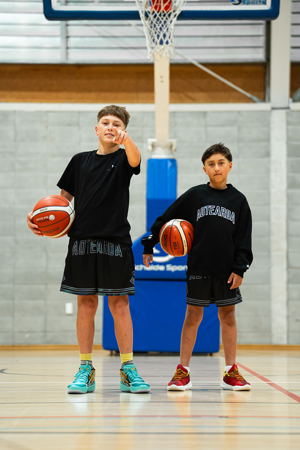Two boys holding basketballs on a basketball court in black aotearoa paua shorts and black aotearoa sweatshirt.