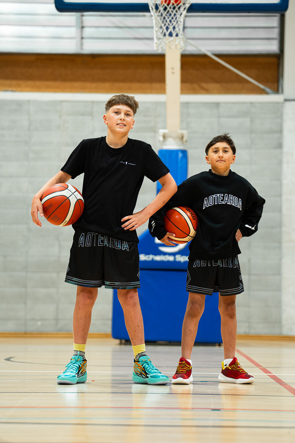 Two young boys holding basketballs on a gymnasium floor in black aotearoa paua shorts