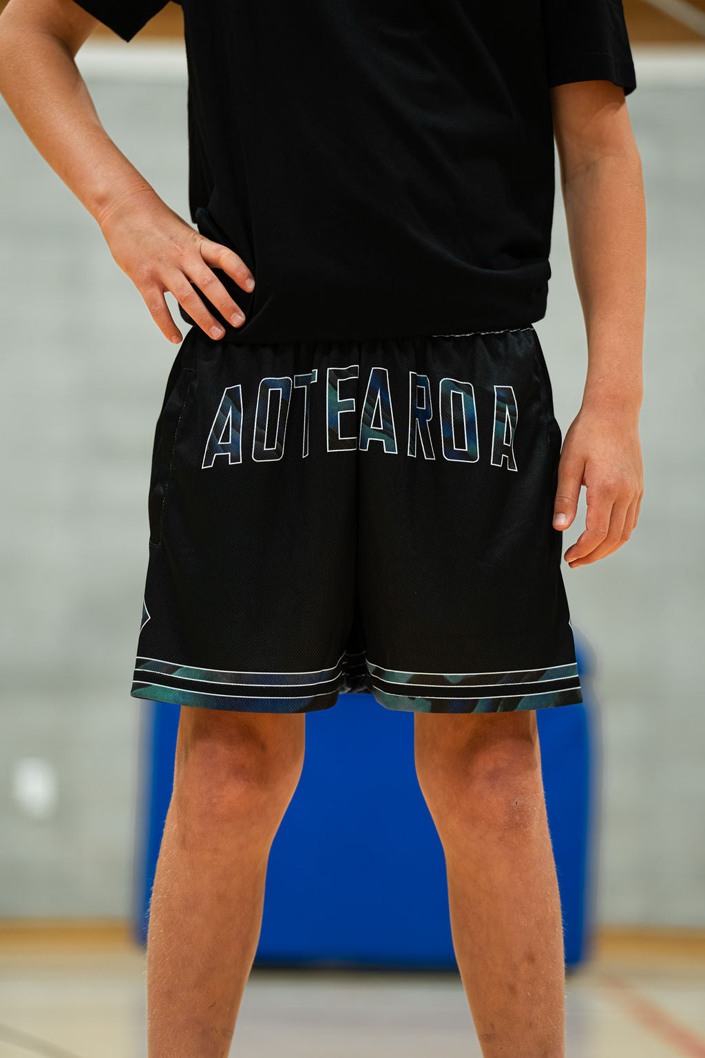 Kid wearing black shorts with 'Aotearoa' on a blurred indoor background