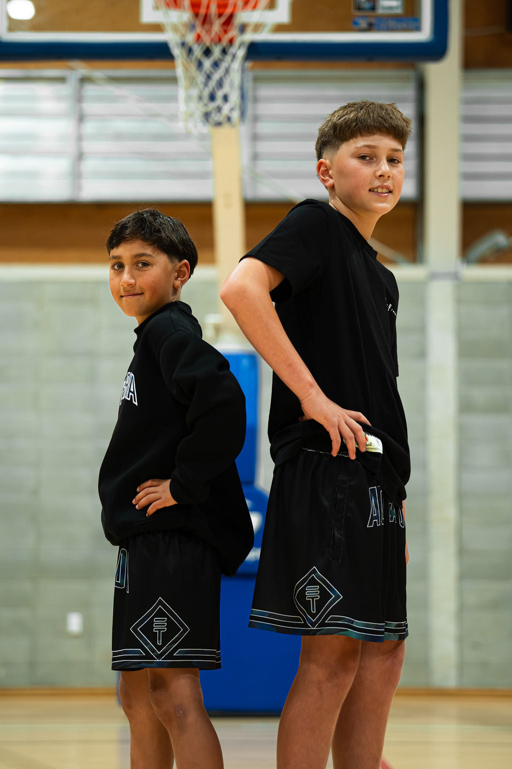 Two boys standing back to back on an indoor basketball court in black kids basketball shorts