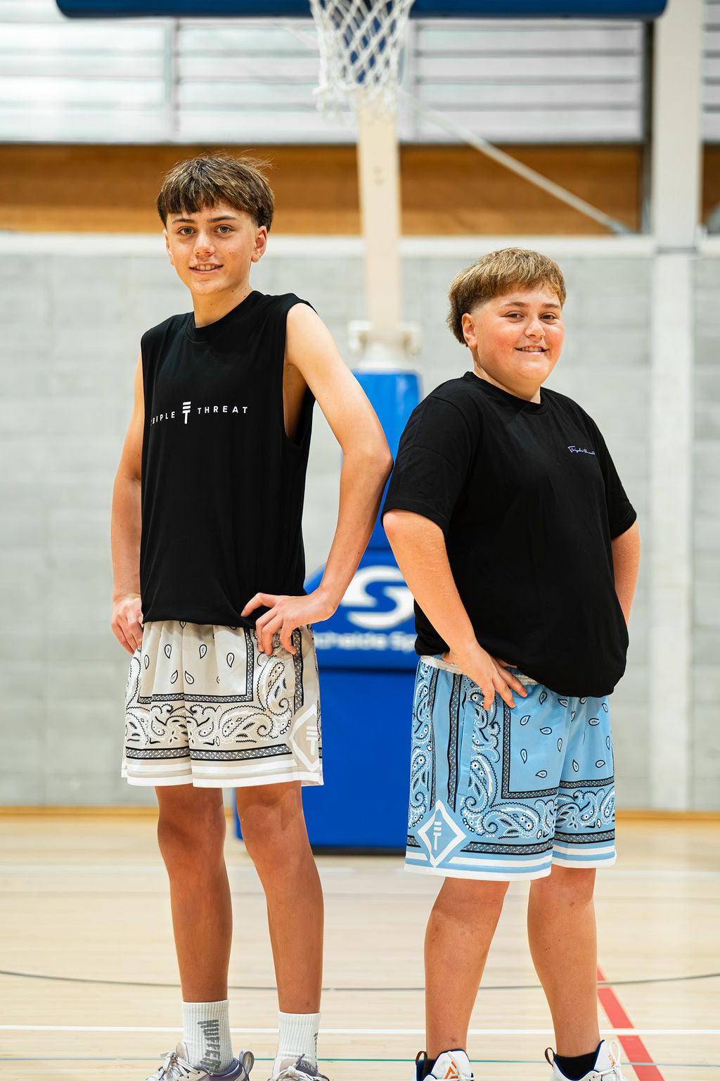 Two young boys standing on a basketball court wearing black t-shirts and paisley patterned kids shorts.