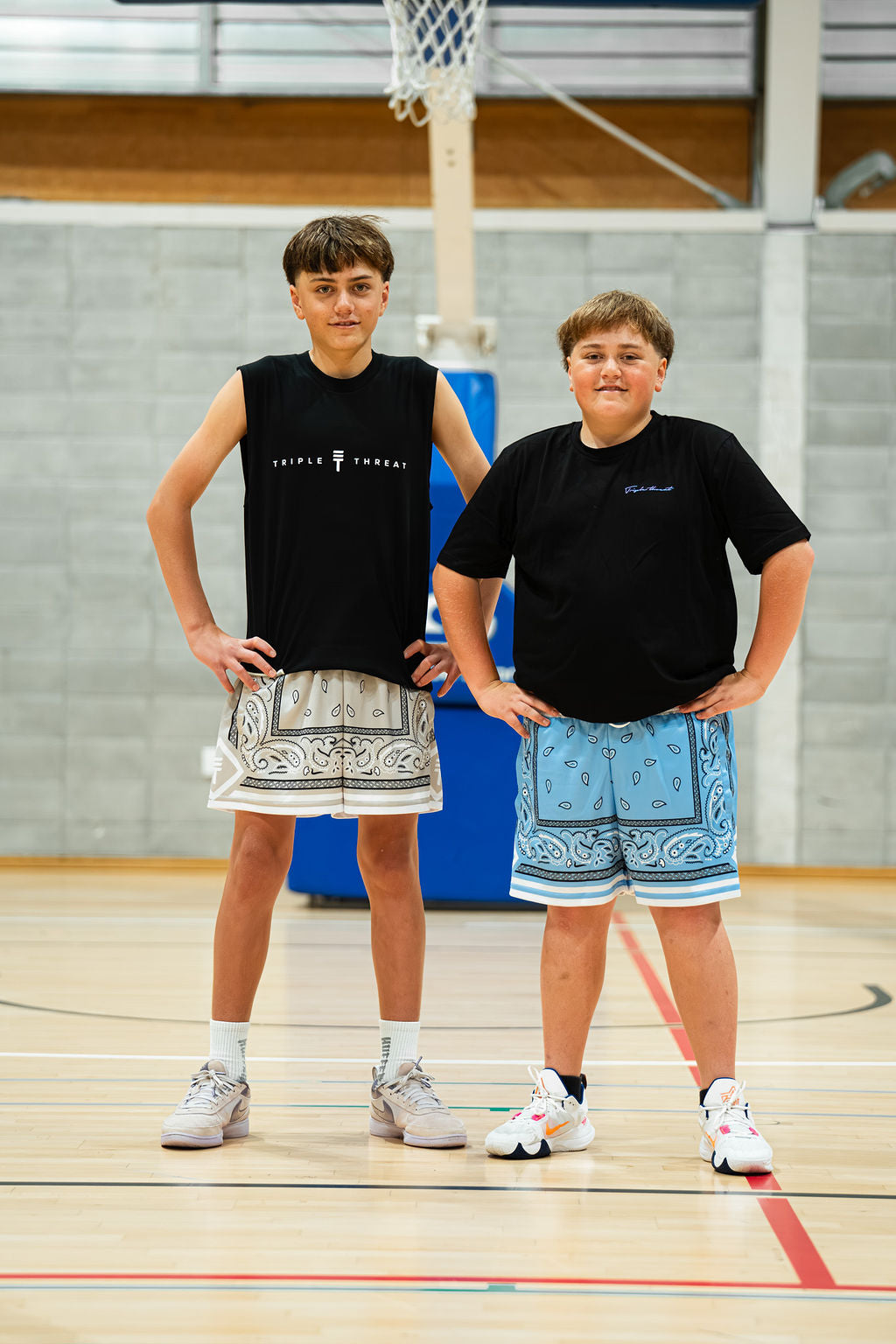 Two young men standing on a basketball court wearing paisley shorts and black t-shirts.