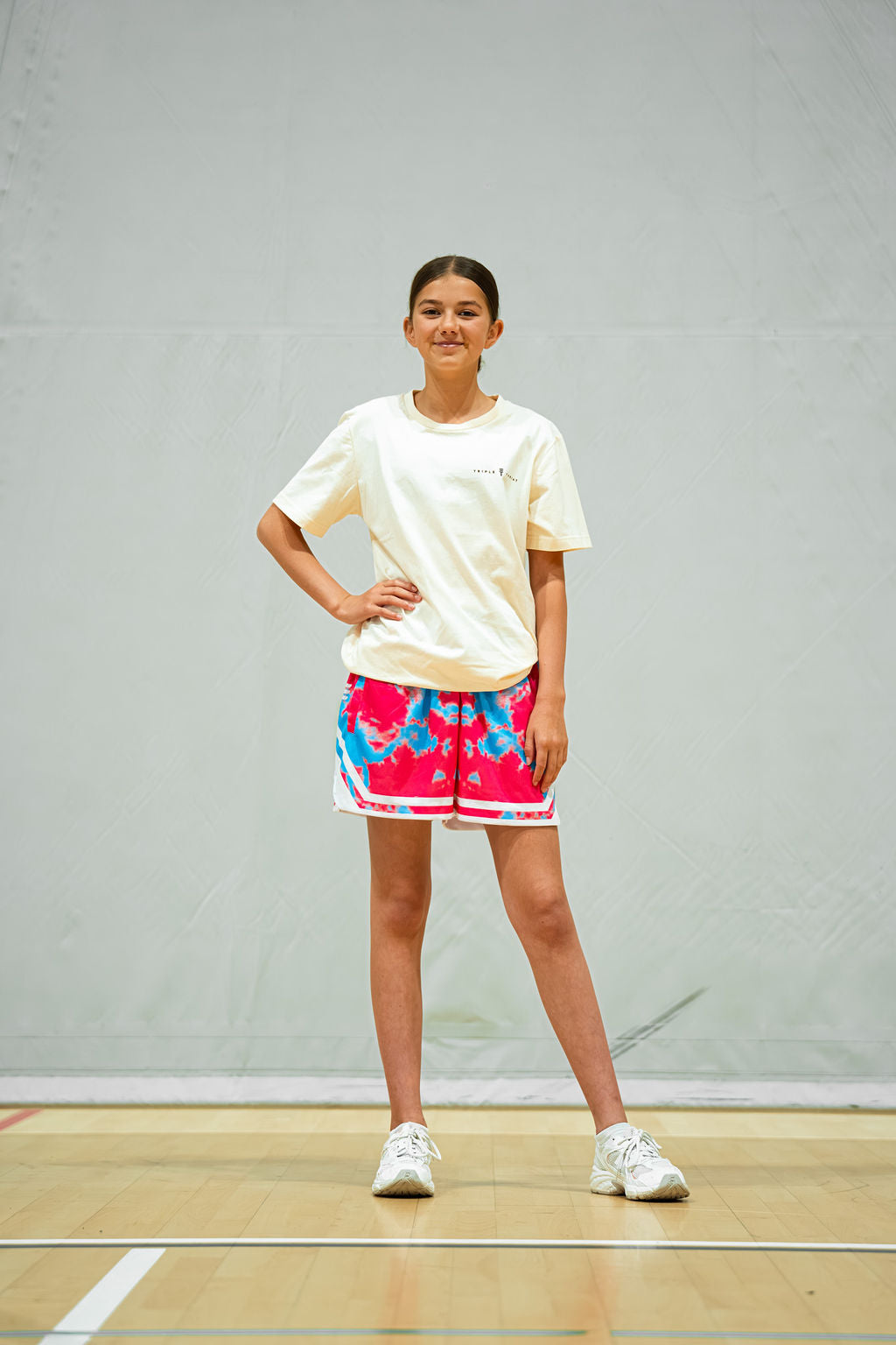 Young girl in a cream t-shirt and colorful shorts standing on a basketball court.