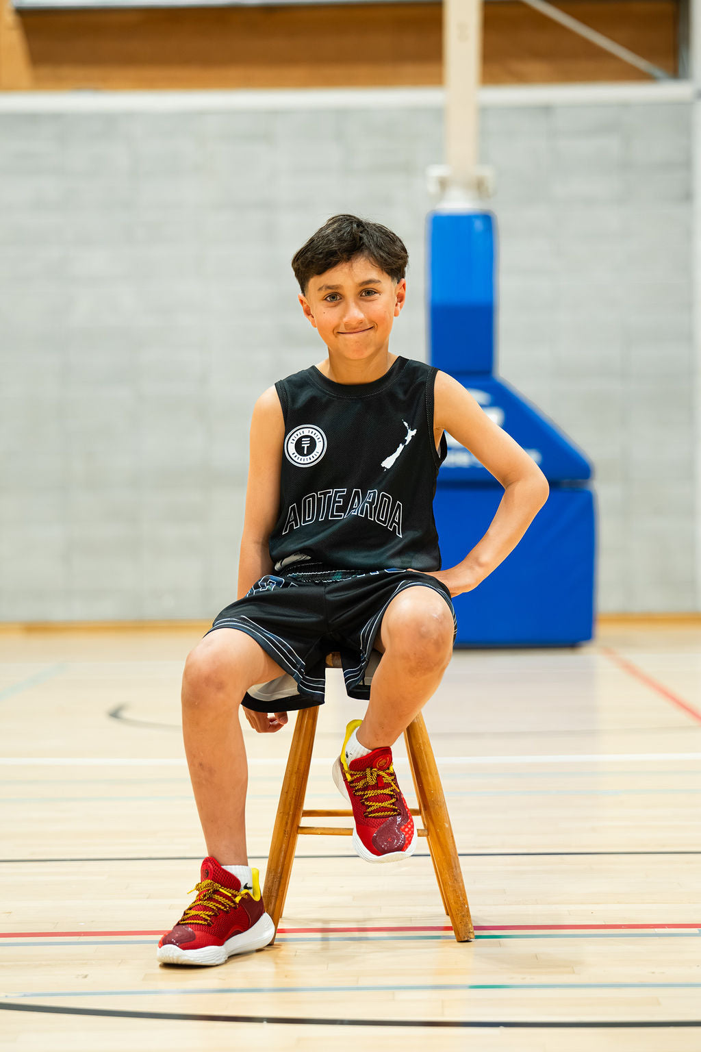 Young boy sitting on a stool in a gymnasium wearing a kids paua singlet in black and black paua shorts.