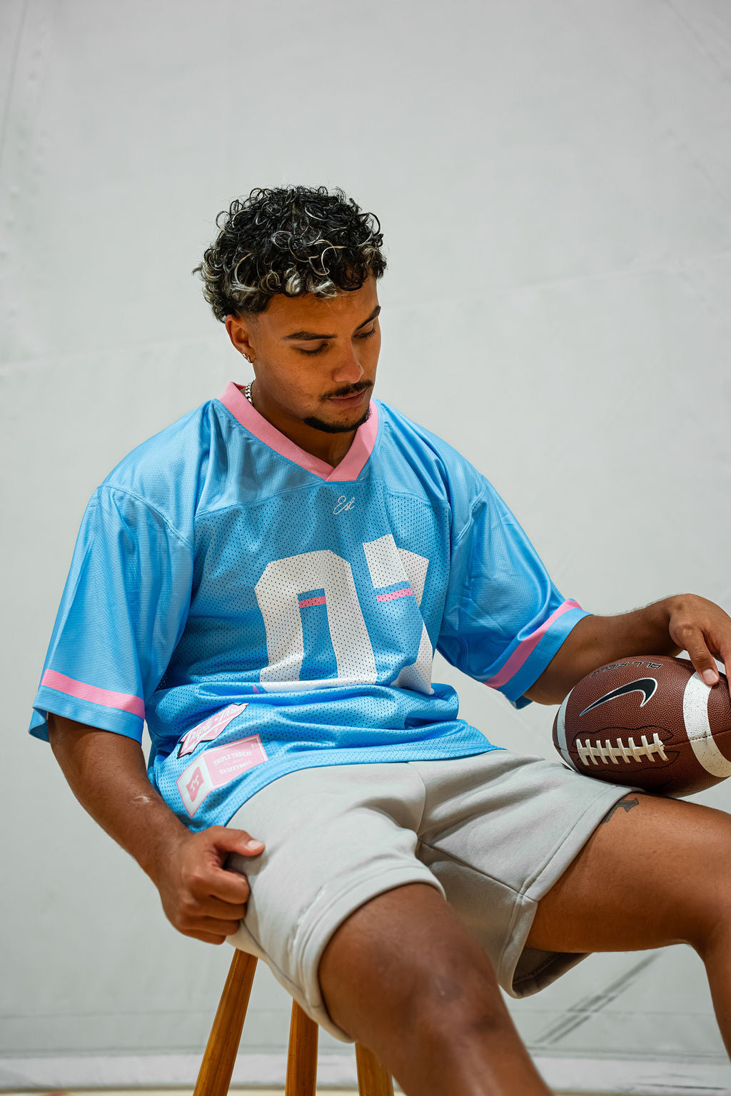 Man sitting on a stool wearing a light blue football jersey with pink accents in light grey shorts, holding a football.