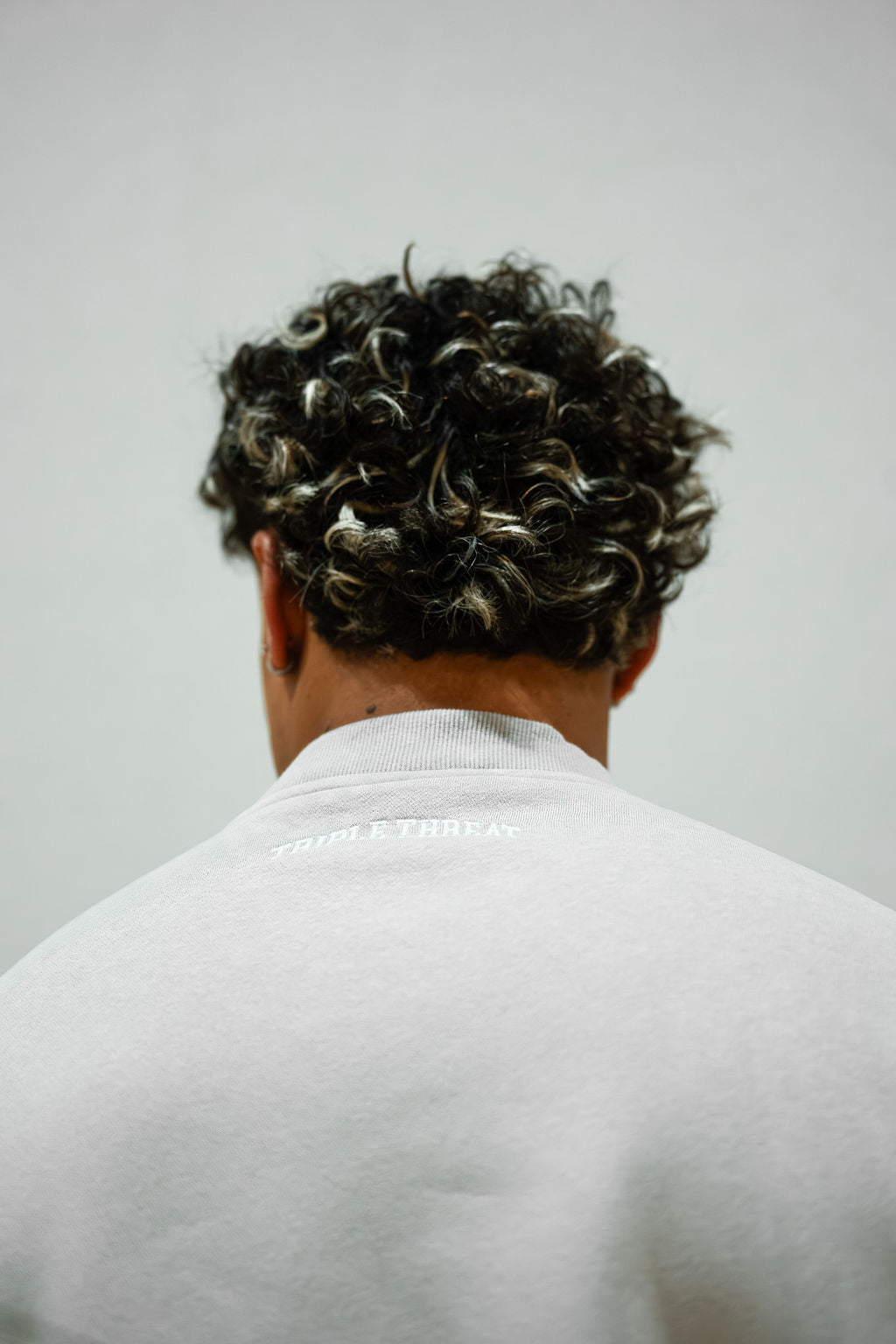 Person with styled hair from behind wearing a light grey crew with Triple Threat embroidered logo on a plain background