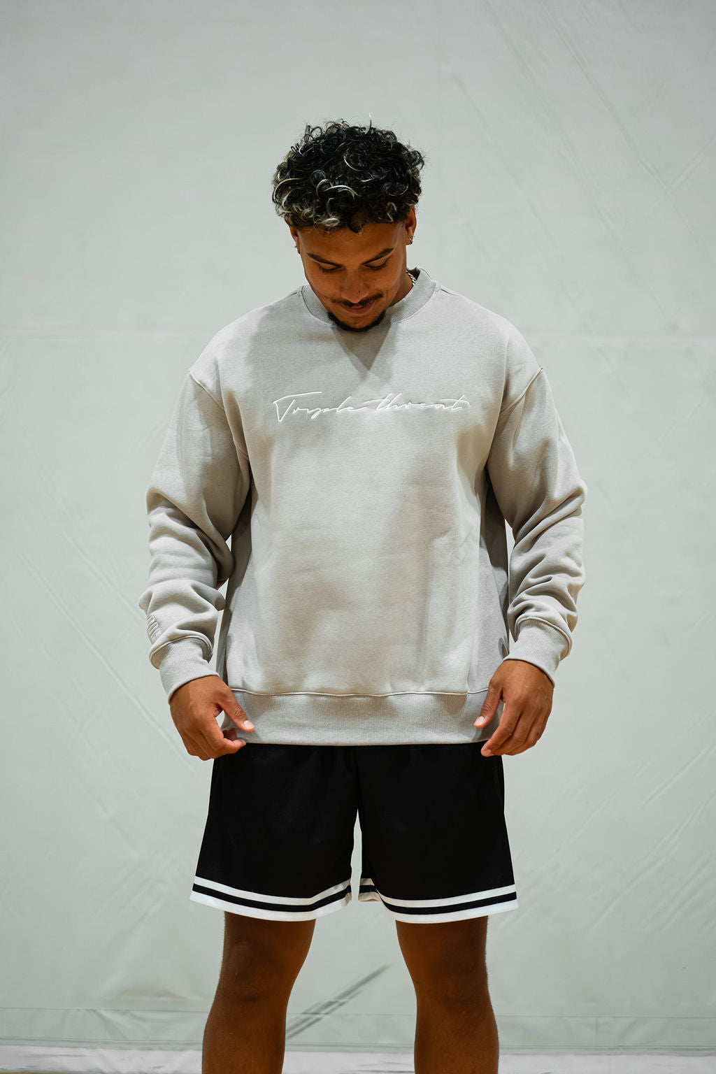 Person wearing a light grey sweatshirt with white embroidered script text and black shorts on a plain background