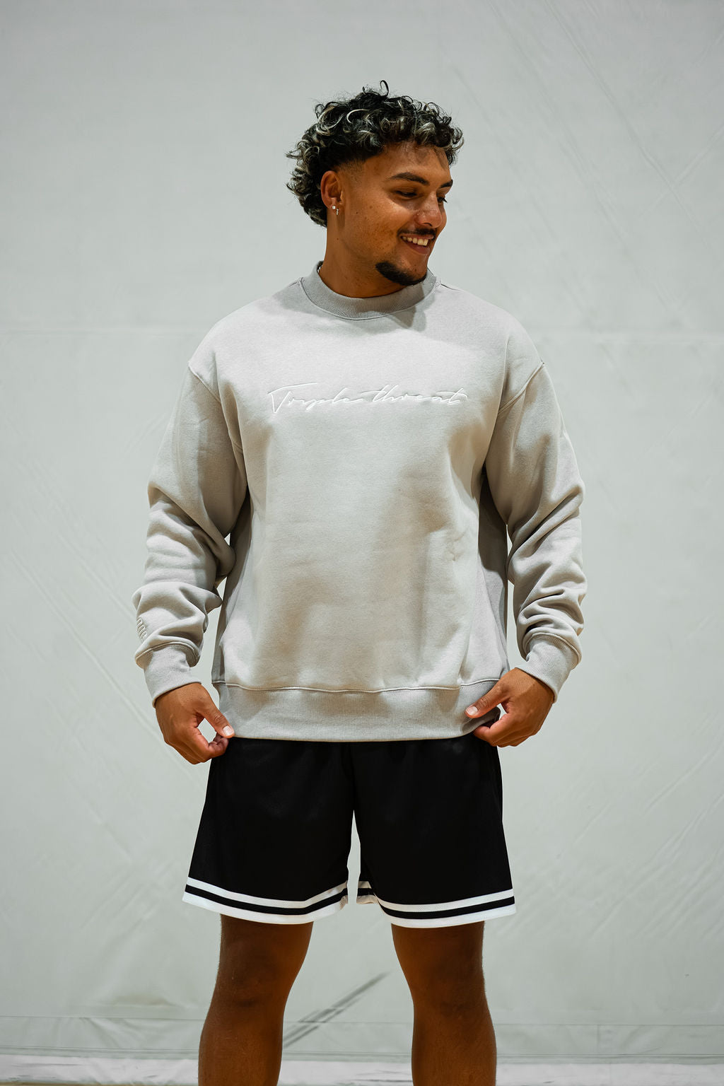 Man wearing a light grey sweatshirt with white embroidered logo and black shorts with white trim against a plain background