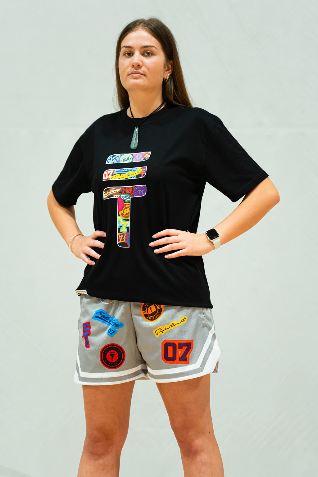 Woman wearing a black t-shirt with colorful graphics and grey shorts with patches on a light grey background