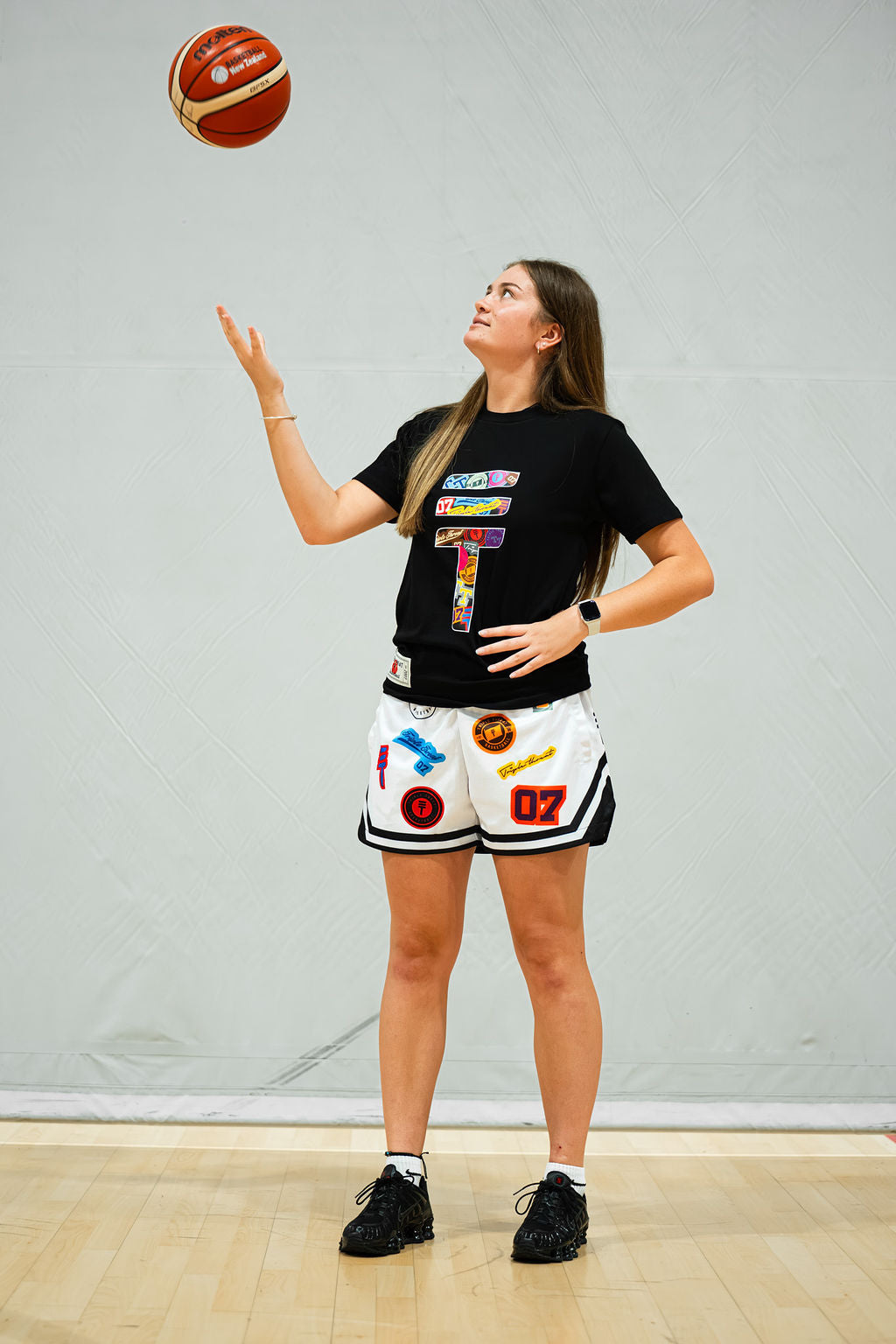 Woman in black sticker bomb t-shirt and white shorts with a basketball, posing against a plain background