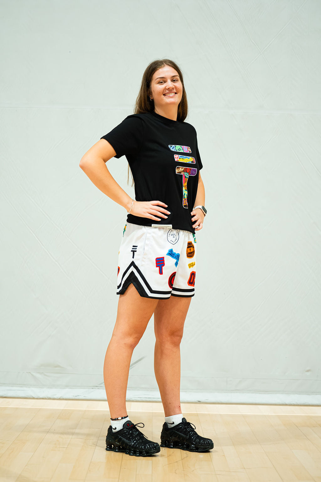 Woman wearing a black t-shirt with colorful graphics and white shorts with sticker bomb logo pattern, standing against a light grey wall.