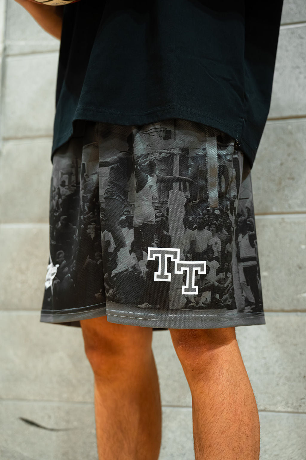 Person wearing black & grey basketball shorts with a pattern of rucker park and 'TT' logo, standing against a stone wall.