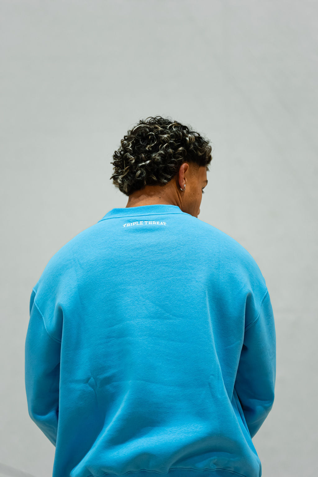 Man wearing a blue crew sweatshirt with Triple Threat logo text on the back against a grey background