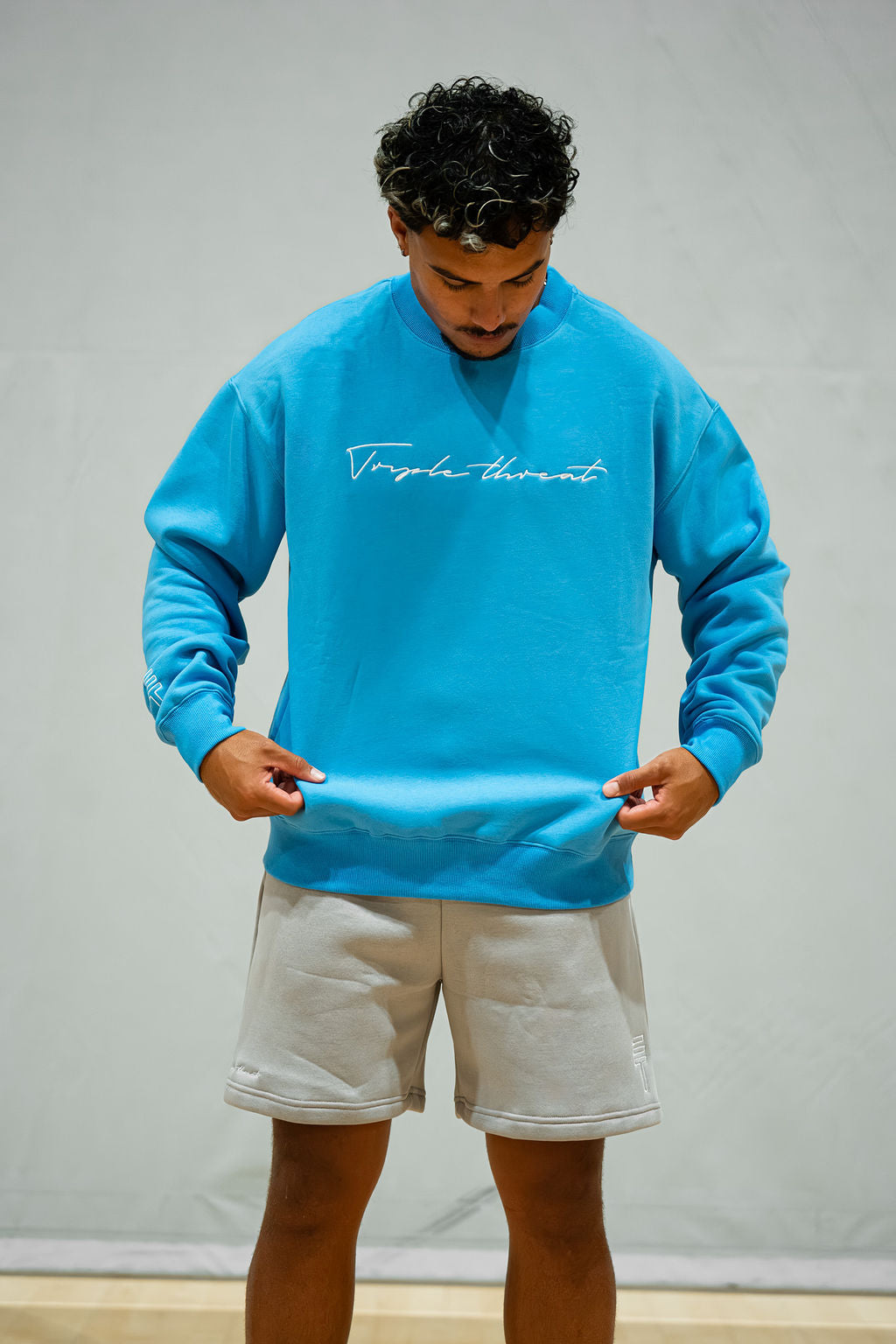 Man wearing a sky blue crewneck sweatshirt with white text and grey shorts against a plain background