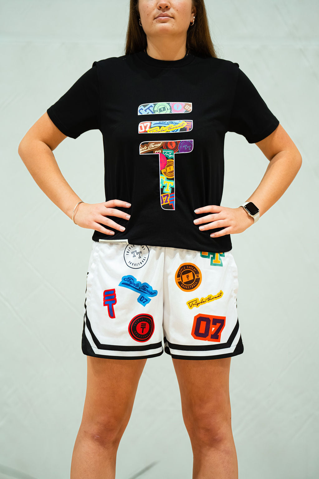 Woman wearing a black t-shirt with colorful graphics and white shorts with various patches on a light grey background