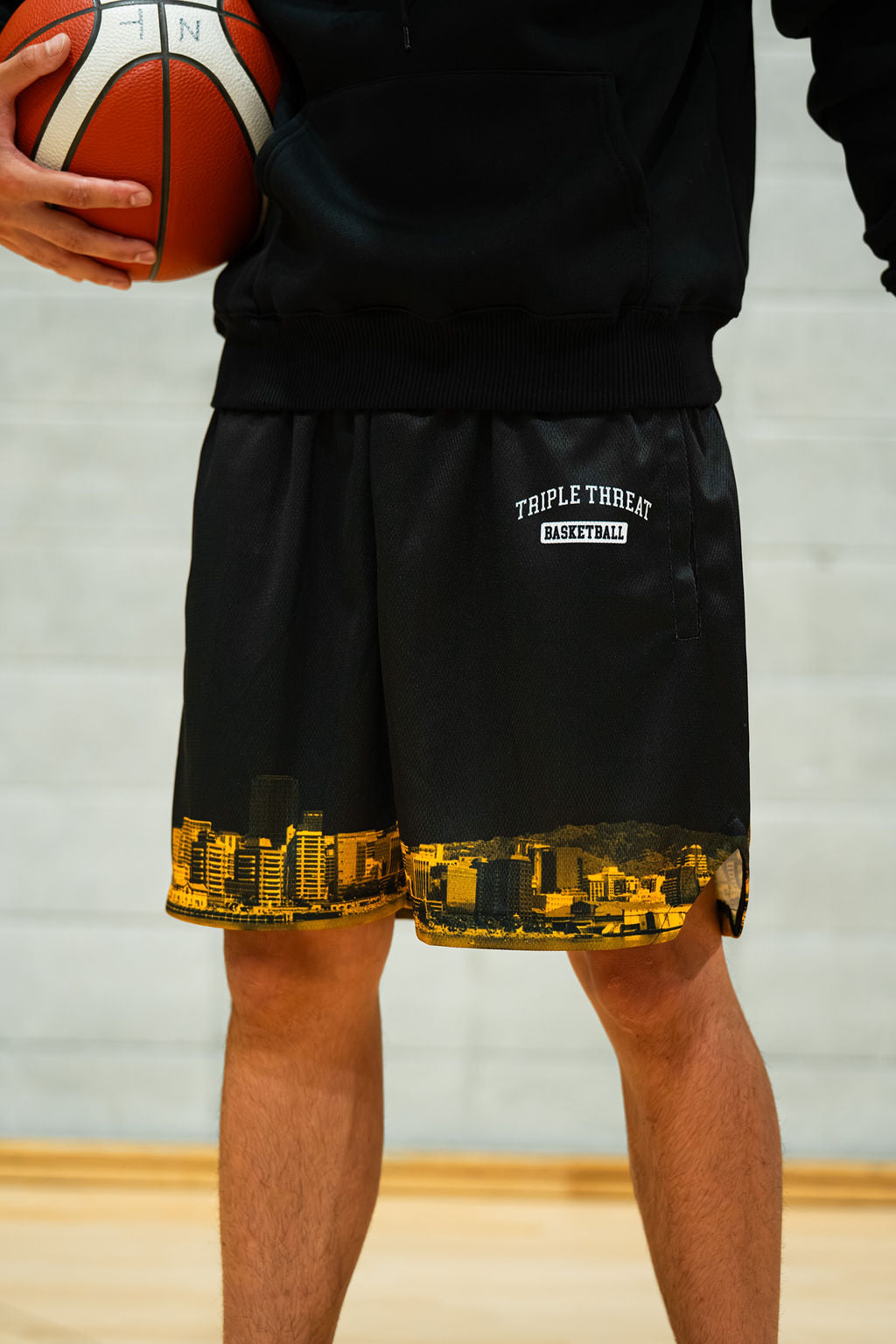 Person wearing black basketball shorts with a yellow cityscape design and 'Triple Threat Basketball' logo, holding a basketball.